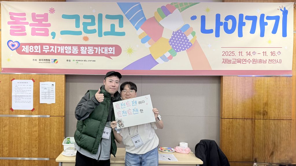 사진 설명 시작. 보석 활동가와 해인 활동가가 제팔회 무지개행동 활동가대회 현수막 앞에 서 있다. 보석 활동가는 엄지를 치켜세우고, 해인 활동가는 트젠트젠하다, 트젠트젠한이라고 적힌 종이를 들어보이고 있다. 사진 설명 끝.